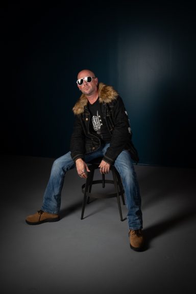 Full body shot of rocker dude with fur collar coat and sunglasses with teal wall and grey floor