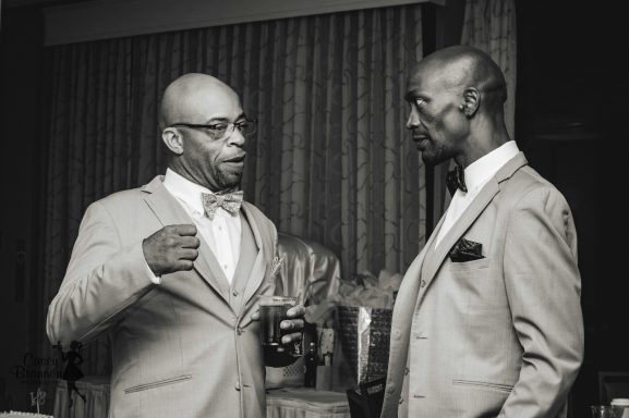 A black and white image of an African American groom and his best man at his wedding talking and having a drink together.