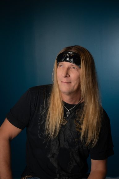 Headshot of long haired rocker looking out of camera with teal background
